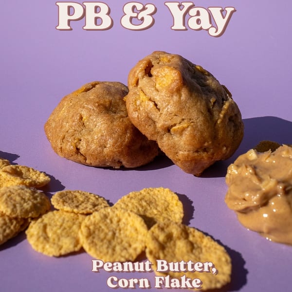 Two peanut butter cornflake cookies on a purple surface with loose cornflakes and peanut butter.