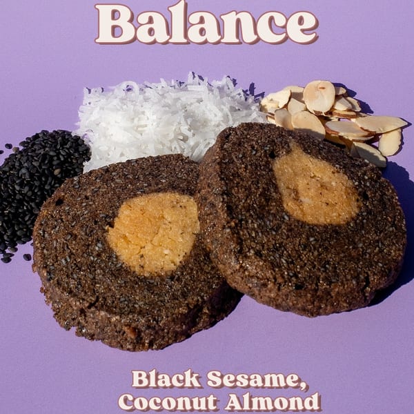 Two black sesame cookies with a light-colored center, surrounded by sesame seeds, shredded coconut, and sliced almonds.