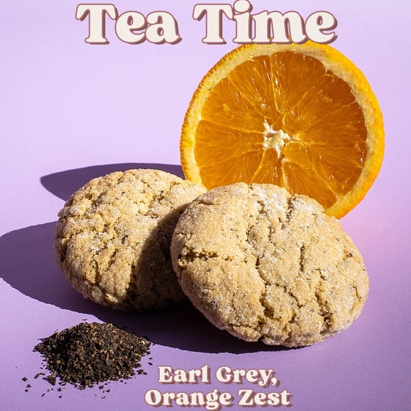 Two Earl Grey orange zest cookies with a sliced orange and loose tea on a purple background.